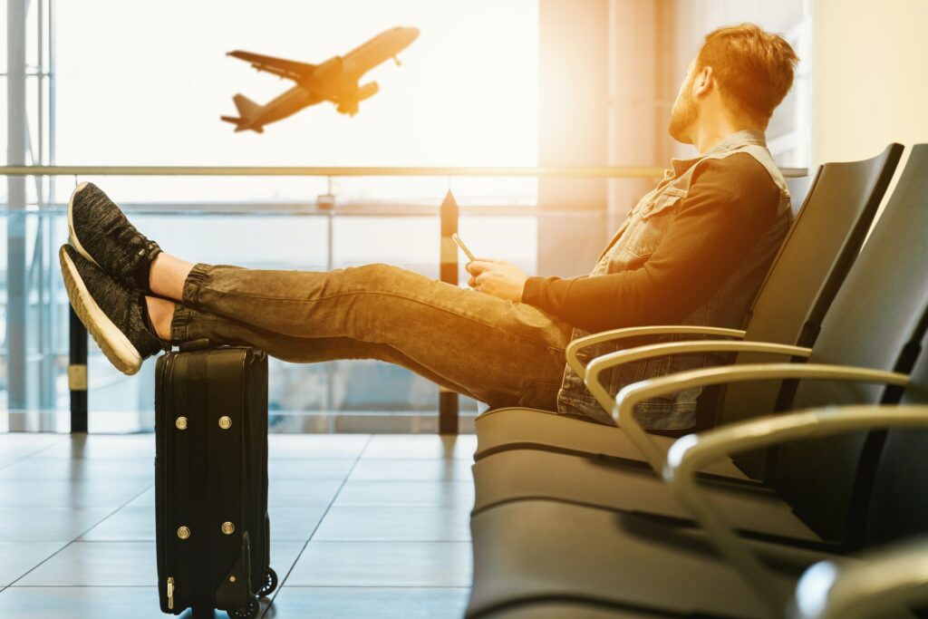 Man sat in airport with feet up on suitcase looking out through window with plane taking off